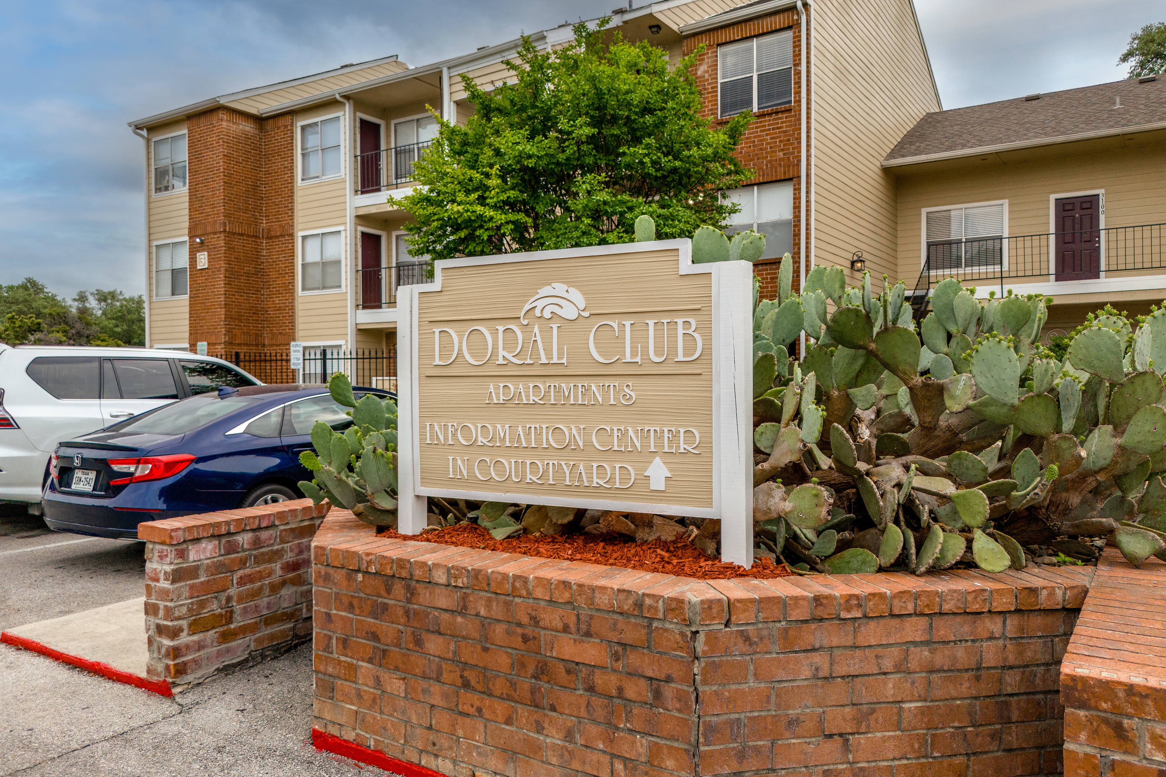 A sign for the Doral Club apartments stands in front of a brick wall and a cactus.